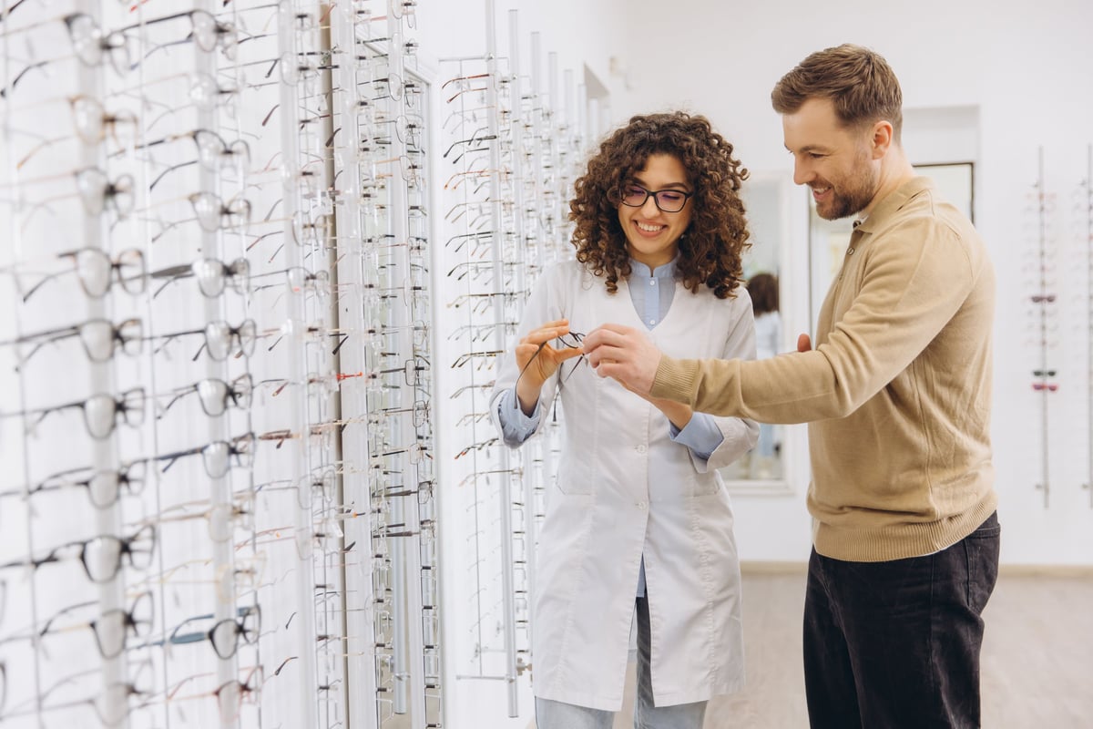 Optician helping man choosing new eyeglasses in optical store, young woman optometrist assisting customer with vision care examination
