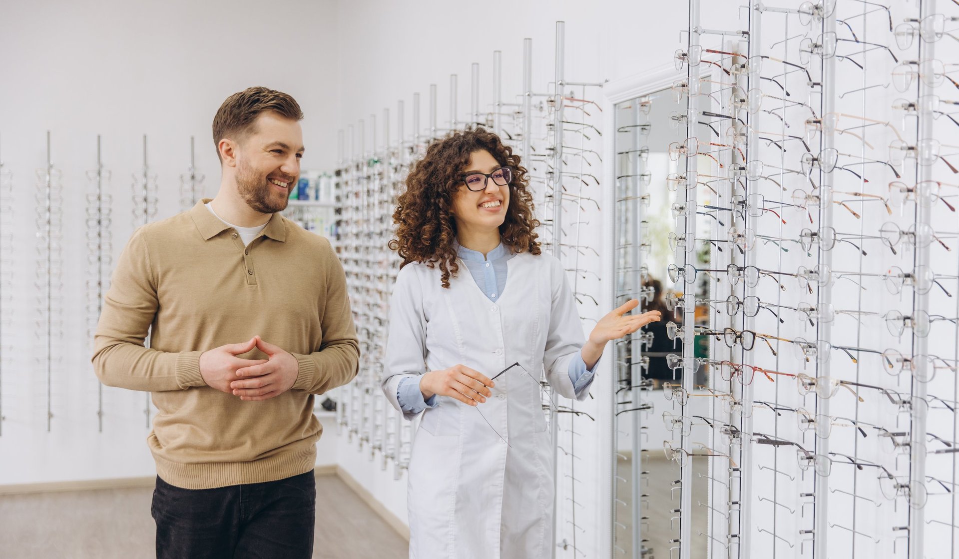Optician assisting a smiling man, showing different eyeglasses frame styles on wall displays in an optometry shop or optical store